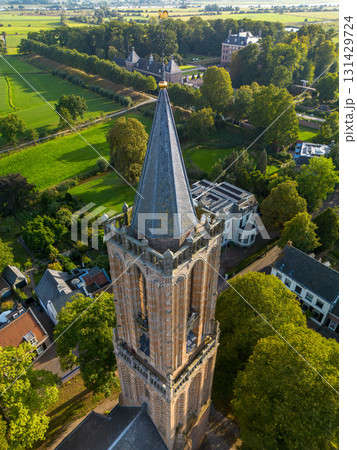 Aerial view of Gothic brick church tower with pointed roof and pinnacles, surrounded by trees and residential houses in a quiet neighborhood. Aerial view of Gothic brick church tower with pointed roof and pinnacles, surrounded by trees and residential houses in a quiet neighborhood. 131429724