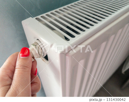 Close-up of a womans hand adjusting a white panel radiator valve at home. Concept of winter heating, temperature control, and energy efficiency. Maintenance action to bleed air, boost heat output 131430042