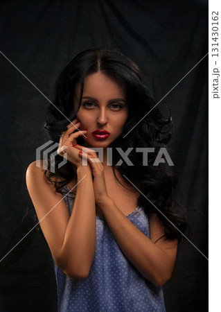 woman stands against a dark backdrop softly illuminated. She wears a simple blue outfit and has long black hair expressing a quiet yet striking mood with her pose and expression. 131430162