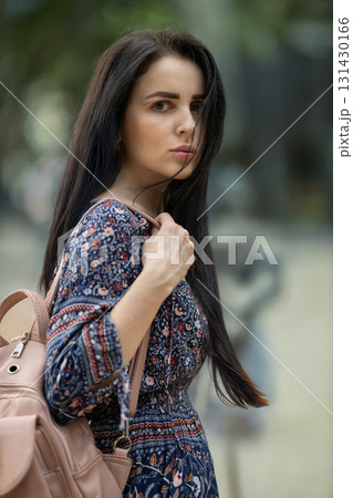 young woman stands outdoors with long dark hair wearing a colorful patterned dress. She holds her backpack and gazes thoughtfully into the distance surrounded by green foliage. young woman stands outdoors with long dark hair wearing a colorful patterned dress. She holds her backpack and gazes thoughtfully into the distance surrounded by green foliage. 131430166