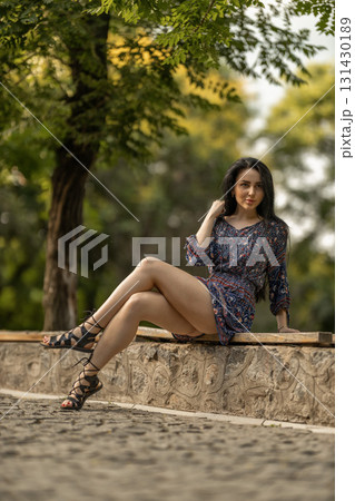 young woman is relaxing on a stone bench in a park. She wears a stylish dress and strappy sandals while the sun shines through trees creating a serene atmosphere perfect for leisure. 131430189