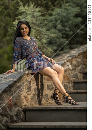 woman with long hair sits casually on stone steps wearing a colorful patterned dress and stylish sandals. Lush greenery surrounds her creating a peaceful atmosphere in the evening. woman with long hair sits casually on stone steps wearing a colorful patterned dress and stylish sandals. Lush greenery surrounds her creating a peaceful atmosphere in the evening. 131430191