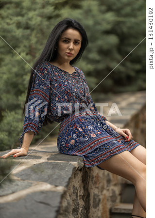 woman sits elegantly on a stone wall wearing a floral patterned dress. Lush greenery surrounds her creating a serene and peaceful atmosphere in the outdoor location. woman sits elegantly on a stone wall wearing a floral patterned dress. Lush greenery surrounds her creating a serene and peaceful atmosphere in the outdoor location. 131430192