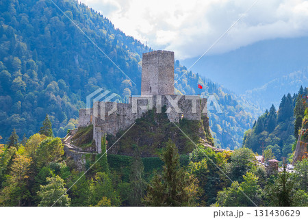 An ancient castle with a stone tower and walls, surrounded by lush greenery and trees, is situated on a rocky hill in a mountainous area. The castle is built with rough stone and has a red flag waving 131430629