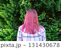 woman with vibrant pink hair stands with her back to the camera, surrounded by lush green foliage of Thuja occidentalis, creating a serene and colorful outdoor atmosphere 131430678