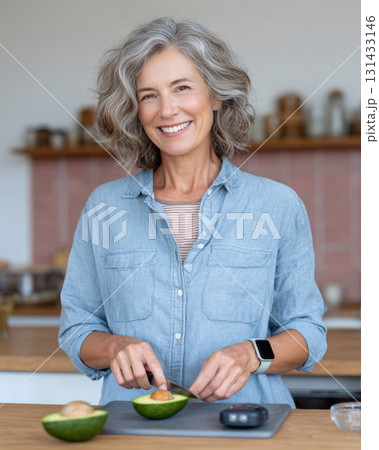 Smiling middle-aged woman slicing avocado in sunlit rustic kitchen Smiling middle-aged woman slicing avocado in sunlit rustic kitchen 131433146