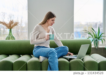 Young relaxing woman with cup looking at laptop screen, sitting on sofa at home 131434520