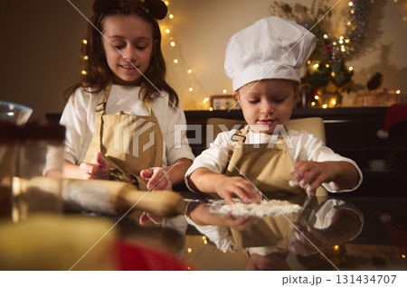 Two Kids Baking Gingerbread Together in Cozy Christmas Kitchen with Aprons and Chef Hat 131434707