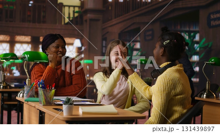 Diverse schoolgirls work with professors on homework next to mockup screen, focused on writing an essay in a school library. Scholars completing their assignment on textbooks. Camera A. 131439897