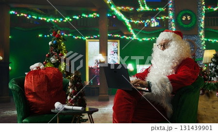 Person dressed as Santa Claus wearing fake beard, red jacket and hat replying back to letters from kids on laptop. Senior man portraying Christmas character answering children using notebook, camera A Person dressed as Santa Claus wearing fake beard, red jacket and hat replying back to letters from kids on laptop. Senior man portraying Christmas character answering children using notebook, camera A 131439901