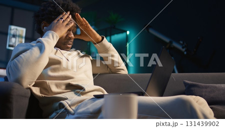 Black person having health problems suffering from headache and nausea, sitting in the living room and struggling to work on tasks. Remote worker suffering from illness at home. Camera B. Black person having health problems suffering from headache and nausea, sitting in the living room and struggling to work on tasks. Remote worker suffering from illness at home. Camera B. 131439902
