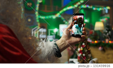 Man dressed as Santa Claus enjoying time off at home watching videos on smartphone. Person wearing Christmas costume spending time online on social media using mobile phone, camera A close up Man dressed as Santa Claus enjoying time off at home watching videos on smartphone. Person wearing Christmas costume spending time online on social media using mobile phone, camera A close up 131439916