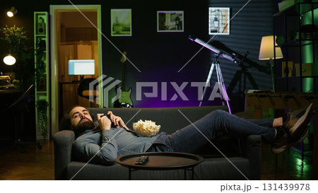 Gamer lounged on couch focused on winning competitive multiplayer game eating popcorn. Person in home theatre holding controller munching on salty snack, playing videogames, camera B Gamer lounged on couch focused on winning competitive multiplayer game eating popcorn. Person in home theatre holding controller munching on salty snack, playing videogames, camera B 131439978