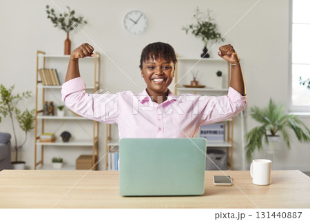 Positive African American woman sitting at desk, raising hands, flexing arms, demonstrating biceps. 131440887