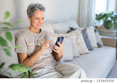 Happy senior woman using smartphone on sofa at home, staying connected and enjoying technology 131441038