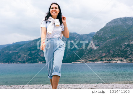 Happy Woman Walking Barefoot on Pebble Beach with Scenic Sea and Mountain View Behind Her 131441106