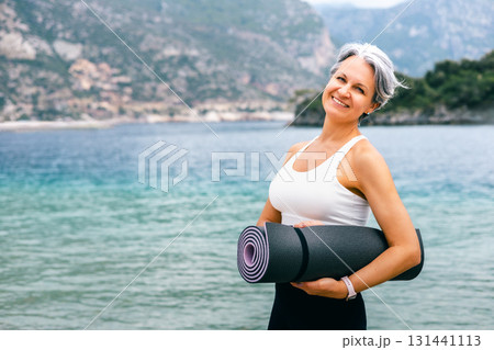Smiling senior woman with yoga mat by the sea, embracing healthy aging and active lifestyle Smiling senior woman with yoga mat by the sea, embracing healthy aging and active lifestyle 131441113