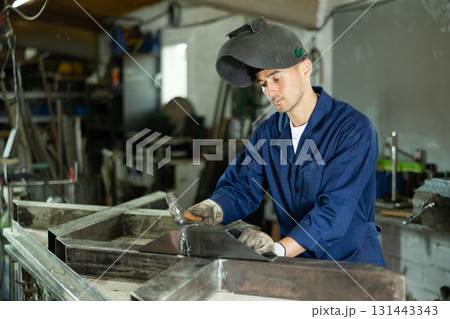 Male welder standing with a welding semi-automatic machine and a safety helmet in metal machining workshop Male welder standing with a welding semi-automatic machine and a safety helmet in metal machining workshop 131443343