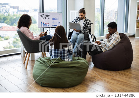 Group of young adults, diverse gender and race, working together in modern casual office. Collaborative atmosphere, bean bags, wheelchair accessibility, and digital devices convey teamwork, inclusion. Group of young adults, diverse gender and race, working together in modern casual office. Collaborative atmosphere, bean bags, wheelchair accessibility, and digital devices convey teamwork, inclusion. 131444880