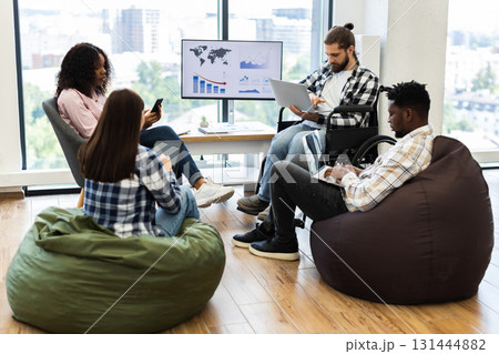 Group of young adults, diverse gender and race, working together in modern casual office. Collaborative atmosphere, bean bags, wheelchair accessibility, and digital devices convey teamwork, inclusion. 131444882