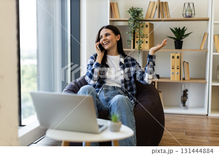Smiling young adult Caucasian woman having phone conversation sitting on bean bag near window Smiling young adult Caucasian woman having phone conversation sitting on bean bag near window 131444884