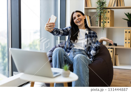 Young adult woman sitting in bean bag chair near large windows, using smartphone for video call while laptop is on nearby table. Indoor setting with bright light and cozy modern decor. 131444887