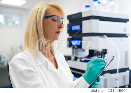 Middle aged blonde scientist analyzing data and writing notes in professional laboratory next to LC MS. Concept of quality research. Middle aged blonde scientist analyzing data and writing notes in professional laboratory next to LC MS. Concept of quality research. 131446944