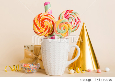 Colorful candies in cup on table on light background background. Large swirled lollipops. Creative concept of a jar full of delicious sweets from the candy store Colorful candies in cup on table on light background background. Large swirled lollipops. Creative concept of a jar full of delicious sweets from the candy store 131449050