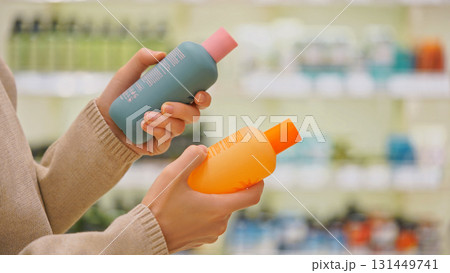 Close-up of female hands comparing cosmetic bottles in a beauty store. Woman reading label and checking ingredients while choosing haircare or body care product. Concept of conscious retail shopping. 131449741