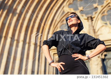 Portrait young adult elegant woman black dress and sunglasses smiles confidently in golden sunlight warm sunset backlit light against historic cathedral building. Female person walking city street 131451302