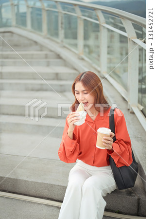 Young woman taking a bite of sandwich while drinking takeaway coffee during a break outside 131452721