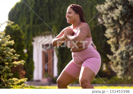 Plus size young woman doing squats on the lawn and looking determined Plus size young woman doing squats on the lawn and looking determined 131453396