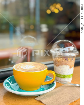 Yellow cup of cappuccino with latte art sits beside a granola yogurt parfait on a wooden table near a cafe window. Concept of coffee, breakfast, healthy lifestyle in a relaxed atmosphere Yellow cup of cappuccino with latte art sits beside a granola yogurt parfait on a wooden table near a cafe window. Concept of coffee, breakfast, healthy lifestyle in a relaxed atmosphere 131454061