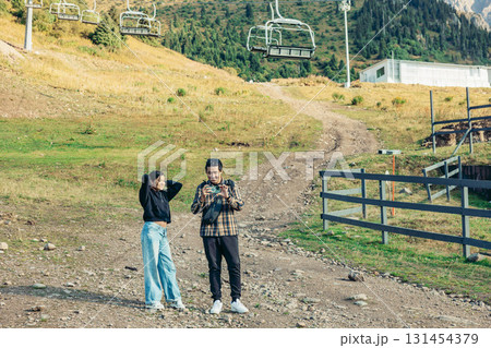 A young Asian woman with long black hair and a young Hispanic man with short black hair stand on a mountain path near a cable car. They are enjoying their vacation. 131454379