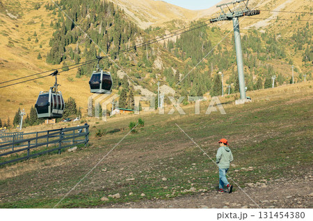 A scenic view of mountains with a cable car in operation. A young child in a red hat walks on the grassy slope, enjoying a vacation in nature. 131454380
