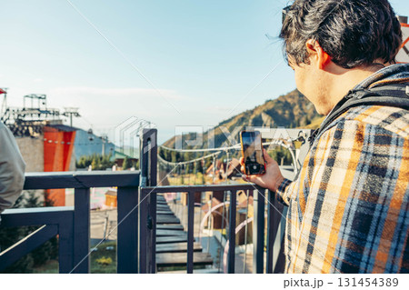 A young Hispanic man with dark hair takes a photo with his smartphone while standing on a balcony overlooking a mountain landscape and recreational area. A young Hispanic man with dark hair takes a photo with his smartphone while standing on a balcony overlooking a mountain landscape and recreational area. 131454389