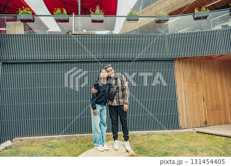 A young Asian woman and a young Hispanic man stand together, smiling in a recreational area with modern architecture and greenery. A young Asian woman and a young Hispanic man stand together, smiling in a recreational area with modern architecture and greenery. 131454405