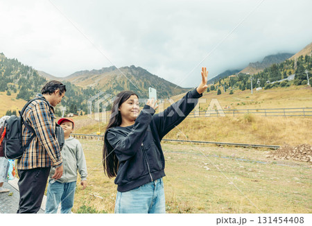 A young Hispanic woman with long black hair takes a selfie in the mountains. Two men and a boy stand nearby, enjoying the scenic view. 131454408