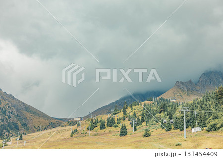 Cloudy mountains with green trees and a ski lift. The landscape features rolling hills and a dramatic sky, ideal for travel and recreation activities. Cloudy mountains with green trees and a ski lift. The landscape features rolling hills and a dramatic sky, ideal for travel and recreation activities. 131454409