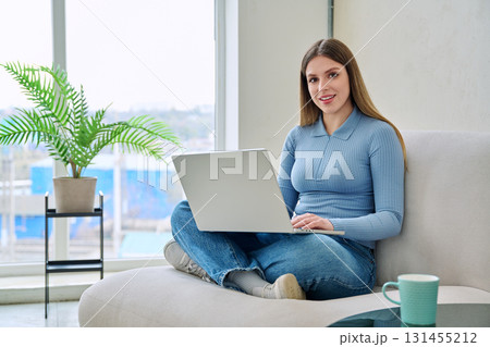 Portrait of young woman sitting on chair at home with laptop looking at camera 131455212