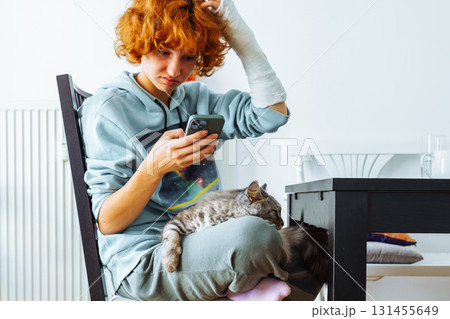 Portrait teenage girl with red hair, cast on arm and domestic cat at home 131455649