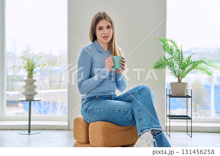 Portrait of relaxed beautiful young woman sitting at home on chair with cup 131456258