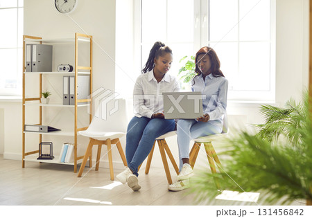 Two African American women sitting on chairs in office and using laptop computer together 131456842
