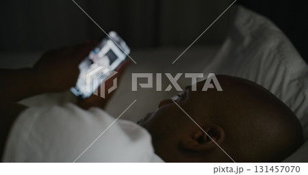Adult Guy Lies in Bed Under Warm Glow of Bedside Lamp, Scrolling Through Social Media on Smartphone 131457070
