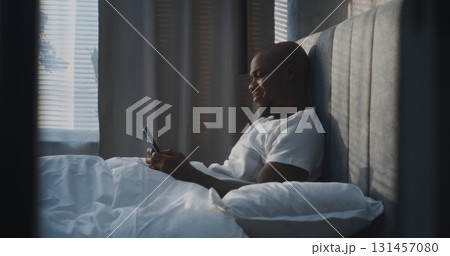 Happy Man Lies Comfortably in Modern Bedroom, Using Smartphone While Leaning on Soft Pillows 131457080