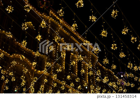 Festive golden lights with snowflake shaped designs hanging on building during night. Sparkling lights create joyful, celebratory atmosphere, perfect for winter or holiday season imagery Festive golden lights with snowflake shaped designs hanging on building during night. Sparkling lights create joyful, celebratory atmosphere, perfect for winter or holiday season imagery 131458314