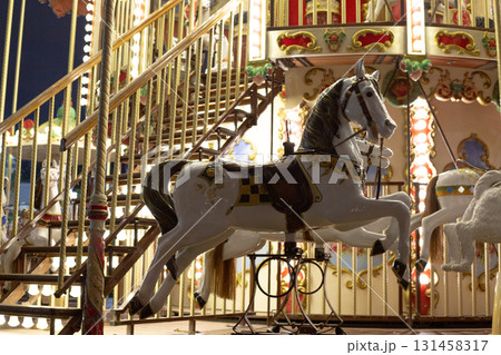 Lit vintage carousel at night, showcasing beautifully painted white horse. The warm lighting highlights the intricate details and nostalgic charm of the fairground attraction 131458317