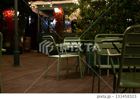 Cozy, dimly lit cafe terrace with metal green chairs and table, surrounded by plants and lights, evokes peaceful, intimate atmosphere perfect for relaxation or quiet conversations under night sky Cozy, dimly lit cafe terrace with metal green chairs and table, surrounded by plants and lights, evokes peaceful, intimate atmosphere perfect for relaxation or quiet conversations under night sky 131458323