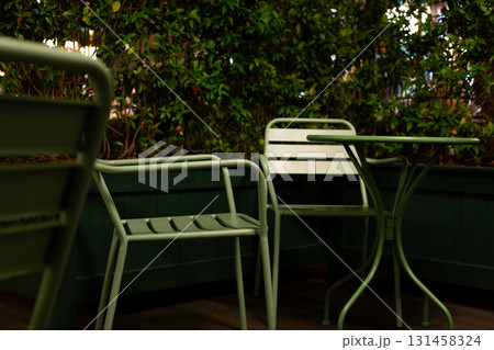Inviting cafe terrace at night, featuring minimalistic green chairs and plants under soft lights, offering quiet escape for those seeking solitude in urban setting with intimate vibe Inviting cafe terrace at night, featuring minimalistic green chairs and plants under soft lights, offering quiet escape for those seeking solitude in urban setting with intimate vibe 131458324