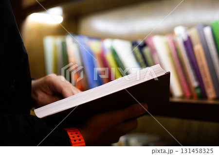 Close up of person holding book in library setting. The photo highlights moment of reading, with shelves full of books in background, creating calm intellectual atmosphere Close up of person holding book in library setting. The photo highlights moment of reading, with shelves full of books in background, creating calm intellectual atmosphere 131458325